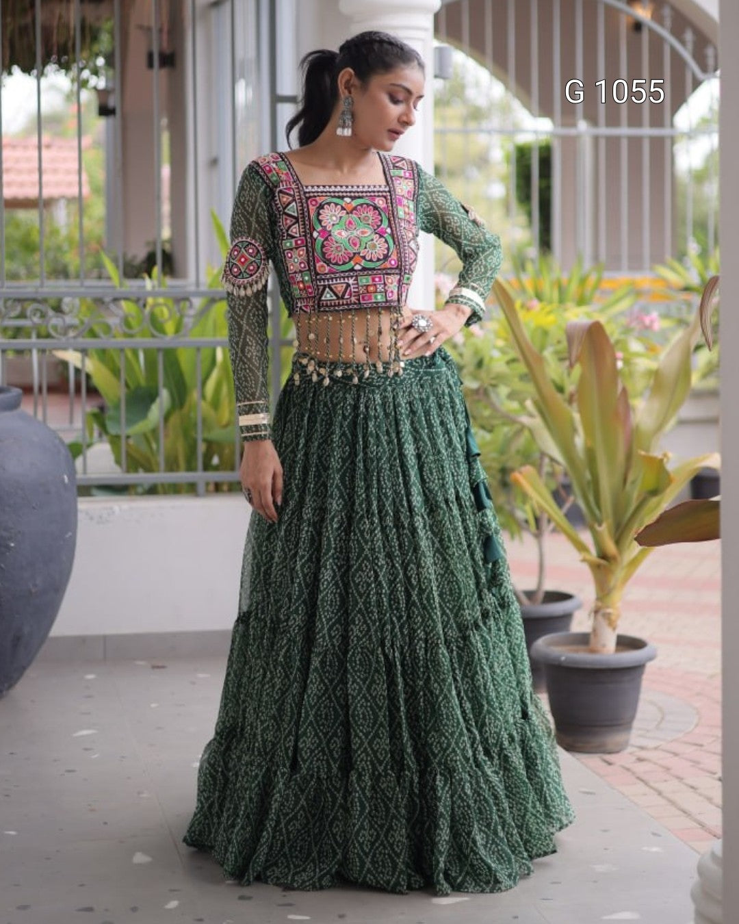 A woman models a traditional Indian outfit consisting of a green embroidered kurta with a matching pleated skirt, standing on a covered walkway with potted plants in the background.