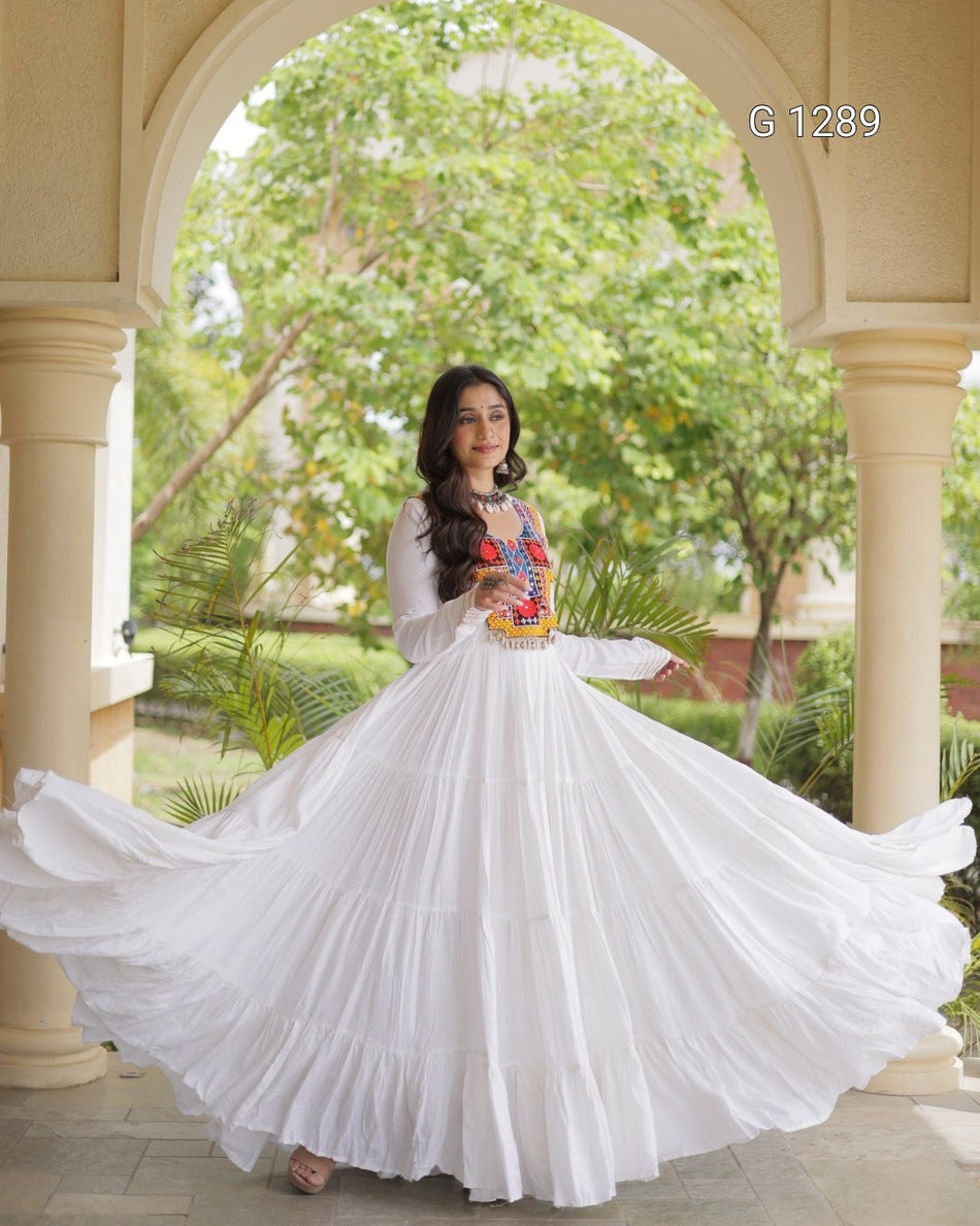 A woman wearing a white tiered long-sleeve dress with a full flowing skirt, standing in a stone archway with green trees in the background. She is holding a small colorful basket.