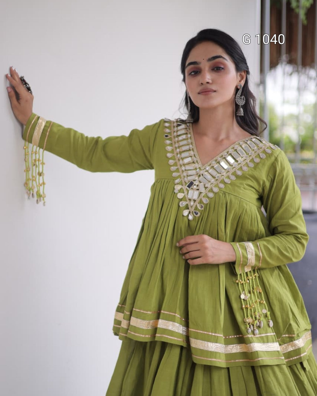 A woman wearing a green traditional kurta with gold mirror work and tassel embellishments, posing against a white wall.