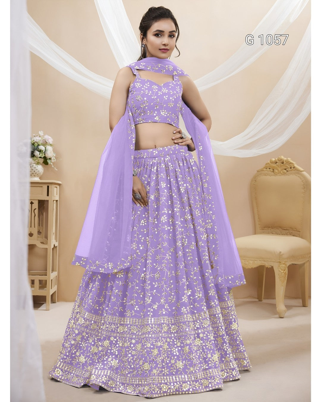 A woman models a lavender purple lehenga choli set with gold floral embroidery and a matching dupatta. The set features a sleeveless crop top, a flared skirt with a detailed border, and a sheer dupatta draped over her arm.