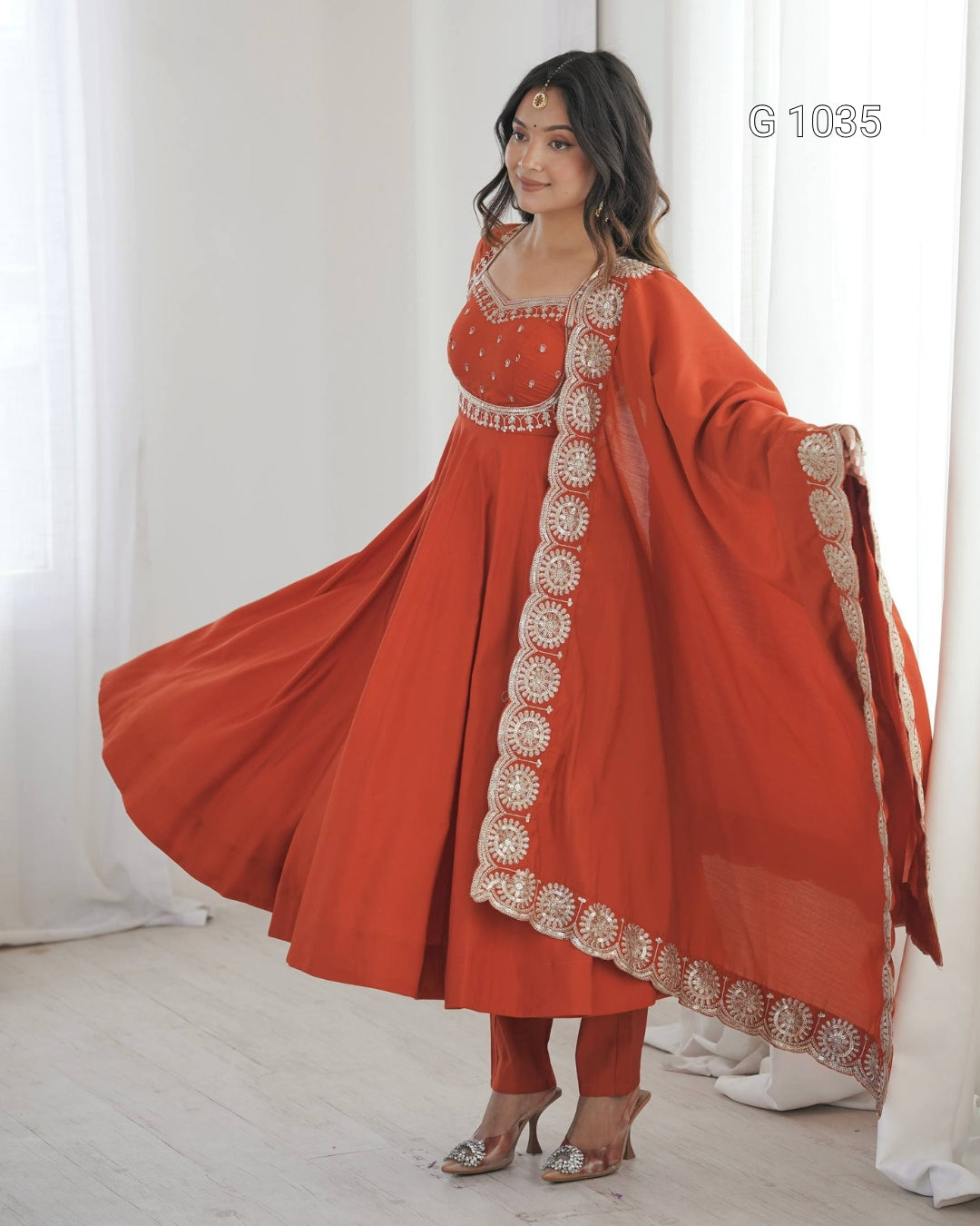 A woman models a traditional Indian outfit consisting of a rust-orange Anarkali kurta with intricate silver embroidery, matching pants, and a matching dupatta with ornate borders, set against a soft white curtain background.