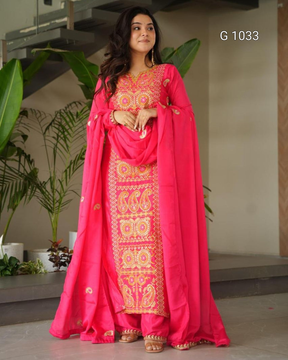 A woman modeling a bright pink traditional Indian salwar kameez suit with intricate gold embroidery, featuring a matching dupatta, in an indoor setting with plants.