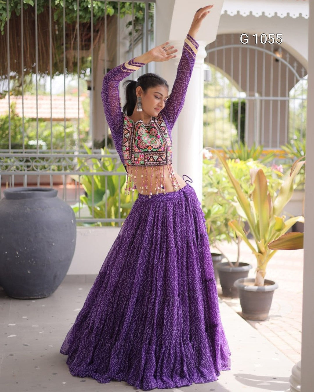 A woman modeling a purple embroidered traditional Indian outfit with a long pleated skirt and a matching embroidered crop top, posed outdoors on a porch with potted plants and a white column in the background.