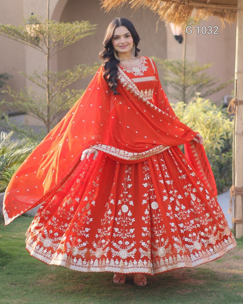 A woman wearing a traditional red and gold embroidered Indian ethnic outfit with a flared skirt and matching dupatta, standing outdoors on grass with trees and a building in the background.