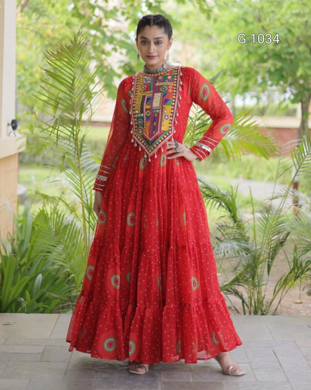 A woman modeling a traditional red long dress with colorful embroidered details on the chest and sleeves, standing outdoors on a tiled patio with green plants in the background.
