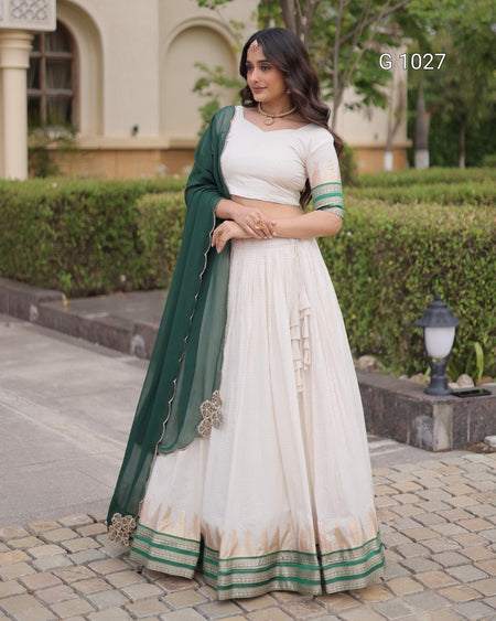 A woman models a traditional Indian ethnic outfit consisting of a cream-colored lehenga skirt, a white blouse with green sleeve borders, and a dark green dupatta draped over her shoulder. The background shows a paved walkway, a green hedge, and a beige building.
