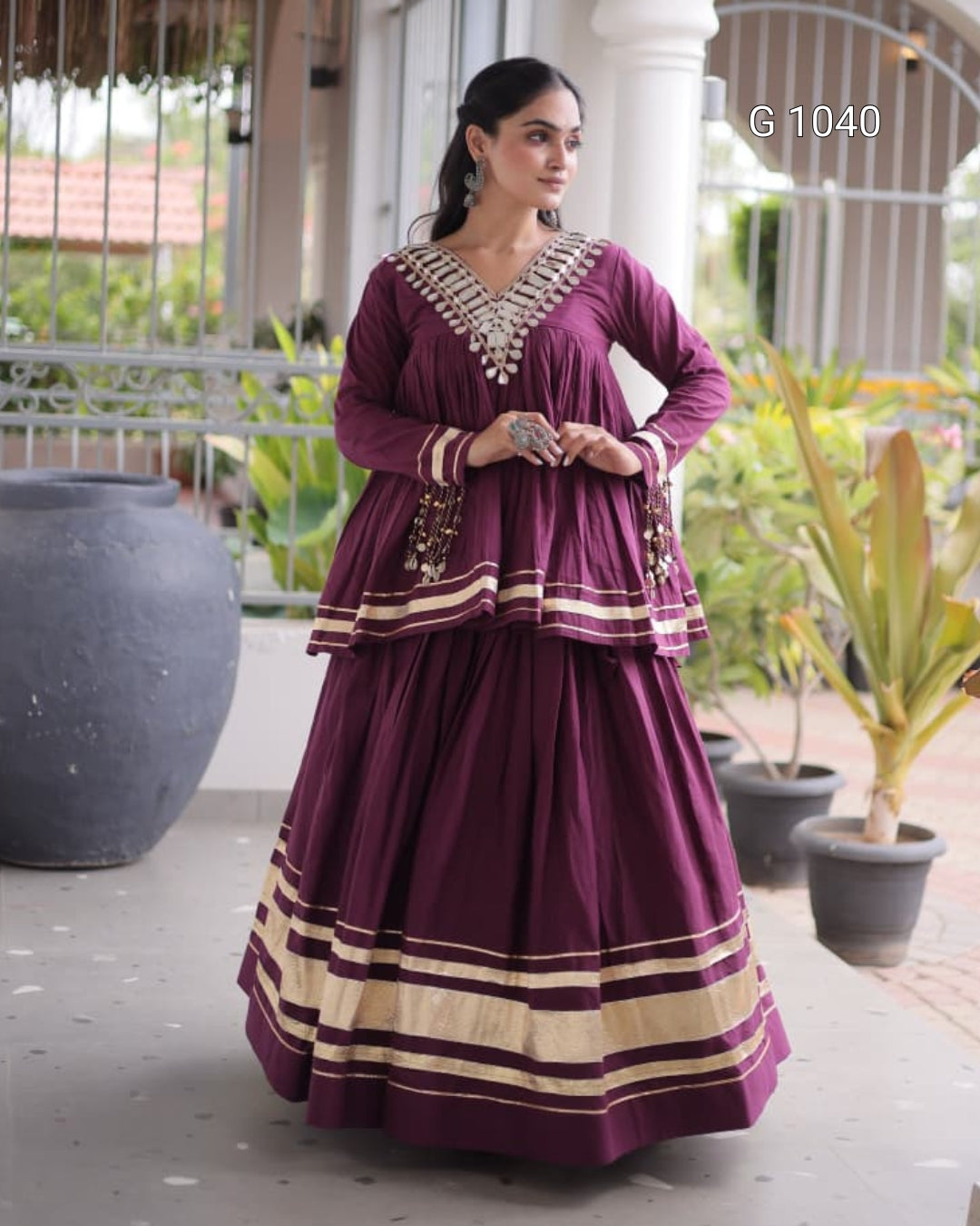 A woman models a traditional Indian outfit consisting of a deep purple long kurta and a matching wide skirt, both decorated with gold stripes and a detailed gold V-neckline. She is standing on a covered porch with potted plants in the background.