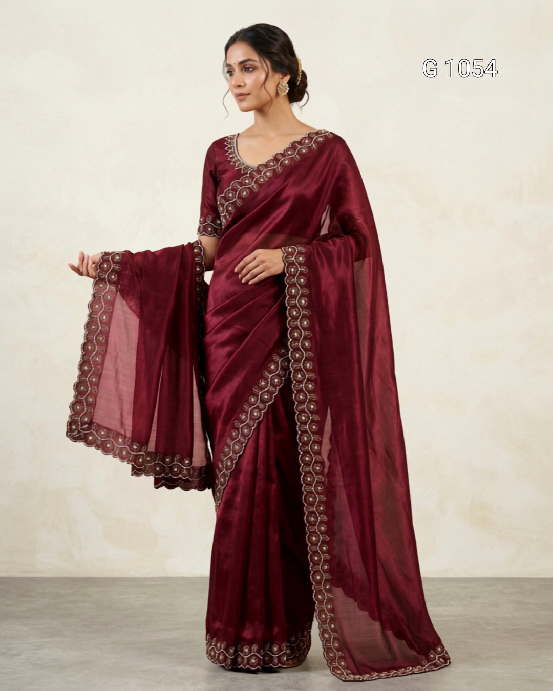 A woman models a deep maroon/burgundy saree with matching blouse and a sheer dupatta, featuring intricate gold and silver embroidered borders. The image is set against a neutral beige textured wall with a gray floor, and the code 'G 1054' is visible in the top right corner.