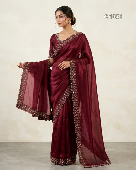 A woman models a deep maroon/burgundy saree with matching blouse and a sheer dupatta, featuring intricate gold and silver embroidered borders. The image is set against a neutral beige textured wall with a gray floor, and the code 'G 1054' is visible in the top right corner.