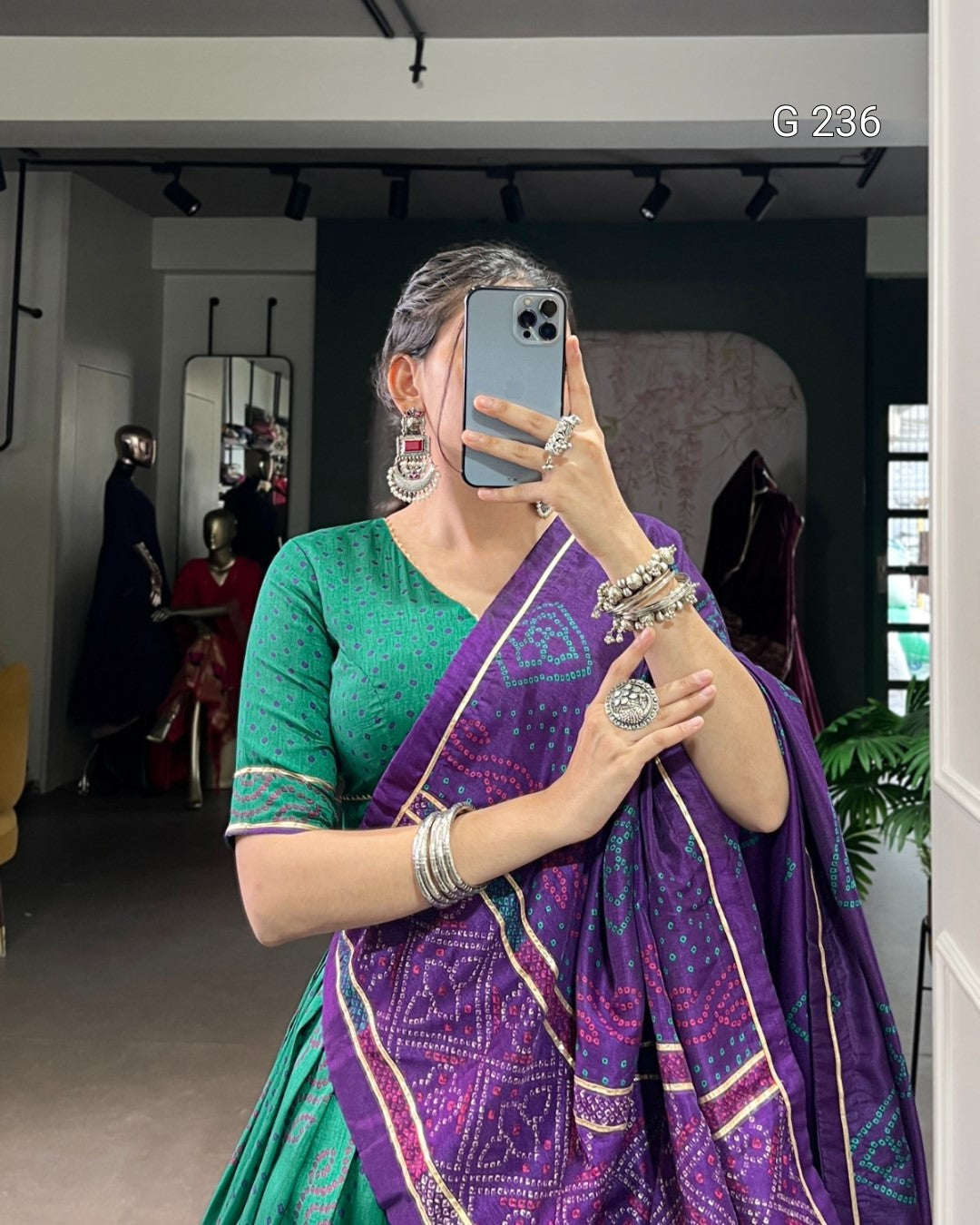 Person wearing a green blouse and purple saree with jewelry, taking a mirror selfie.