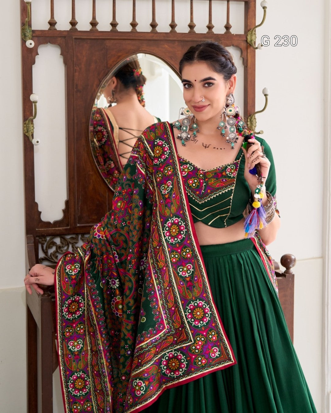 Woman in traditional green and multicolored outfit with jewelry, standing in front of a mirror.