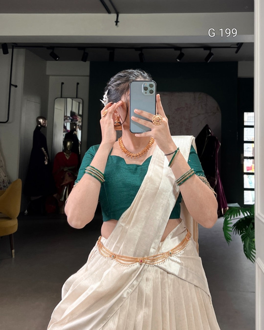 Person wearing a green blouse and white saree with gold border, taking a mirror selfie.