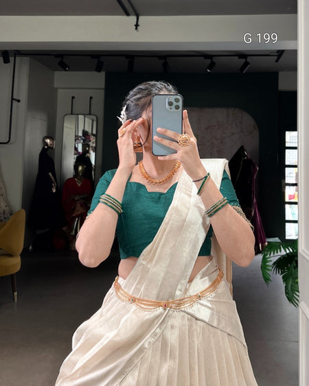 Person wearing a green blouse and white saree with gold border, taking a mirror selfie.