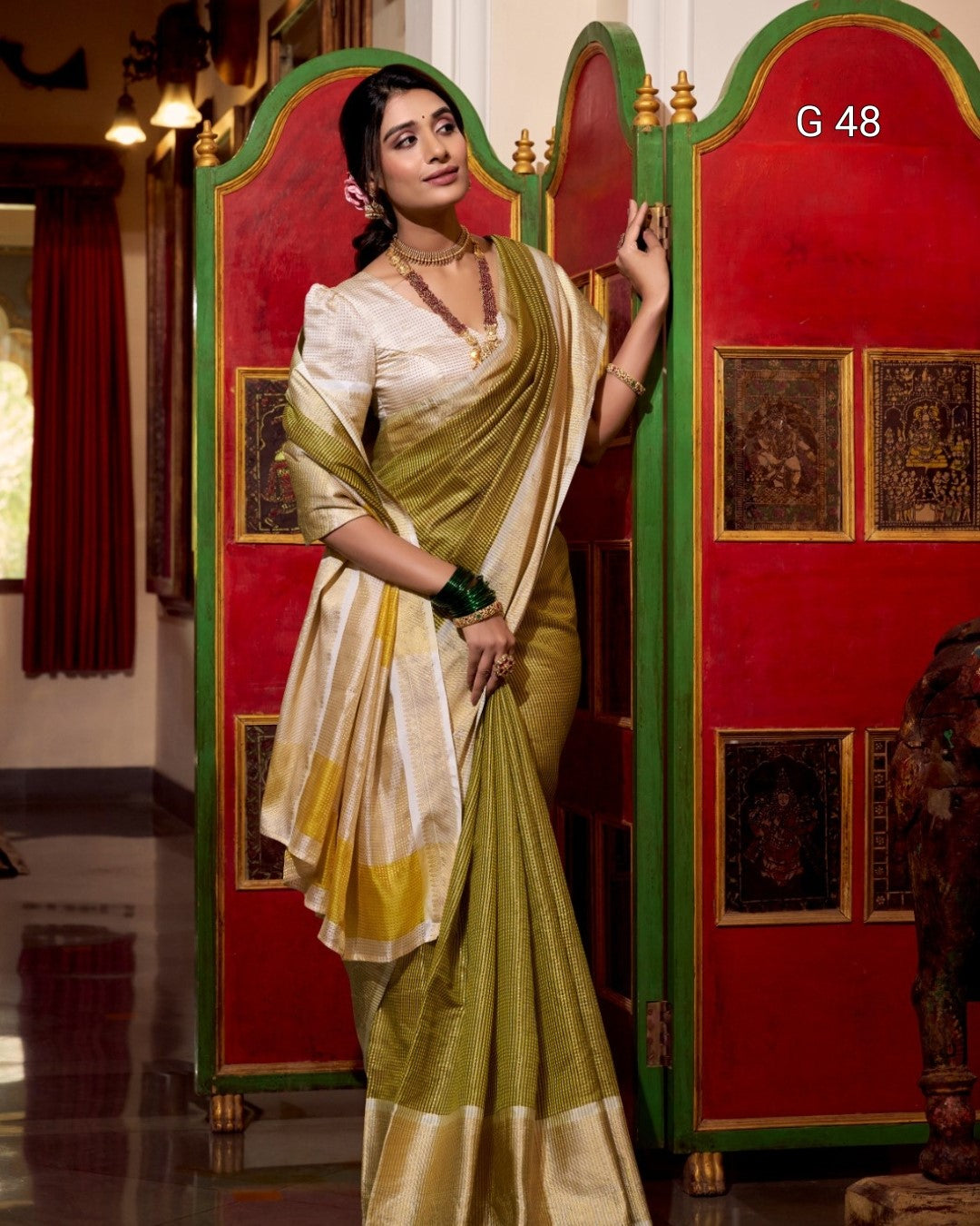 Woman in a green and white saree standing in front of a red and gold decorated wall.