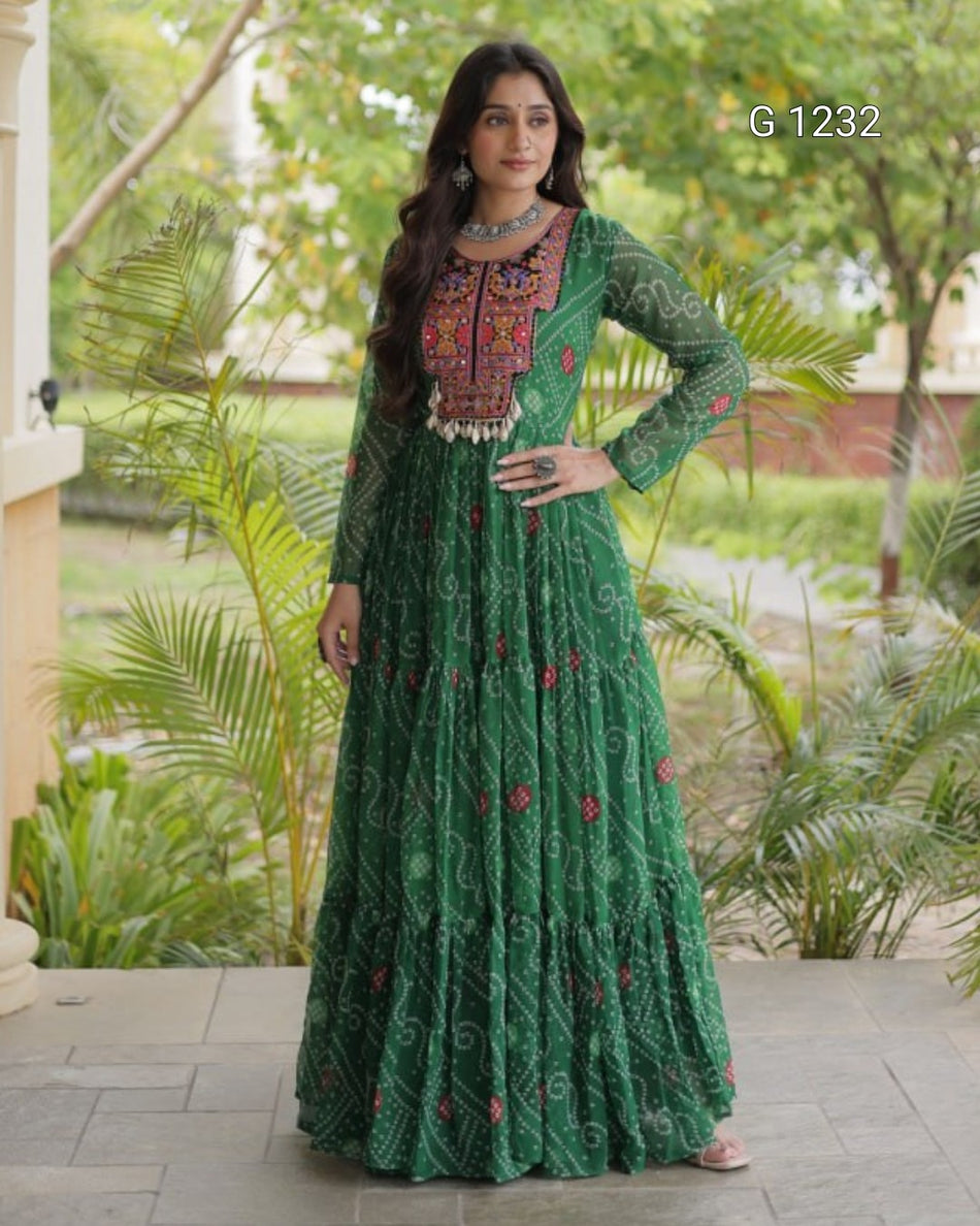 A woman modeling a long green embroidered Anarkali gown with a heavily embellished neckline and red floral motifs, standing outdoors on a paved walkway with green plants in the background.