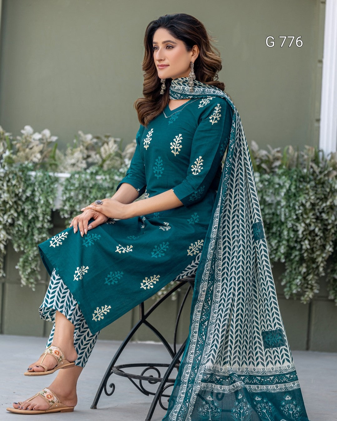 Woman in teal embroidered outfit sitting outdoors with floral background