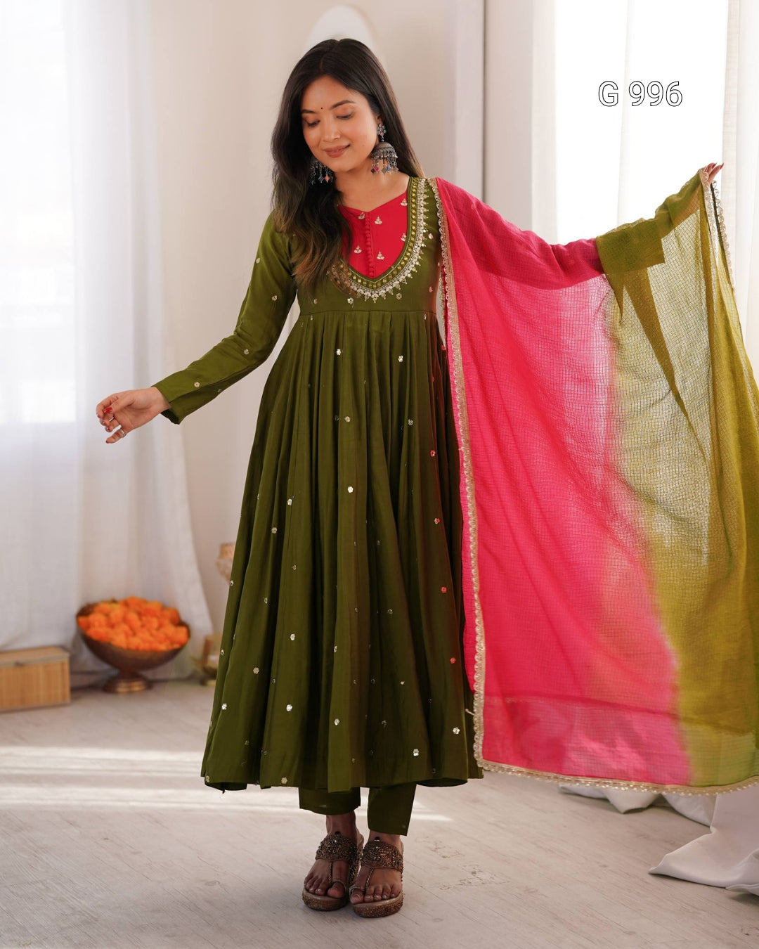 A woman modeling a traditional Indian outfit featuring an olive green long kurta with gold embroidery and a bright pink dupatta with a yellow-green border, set against a softly lit indoor background.