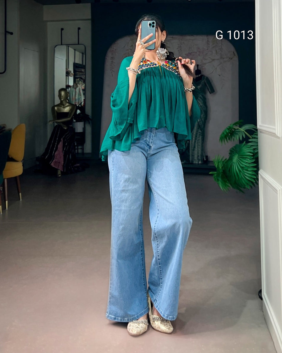 A woman takes a mirror selfie in a boutique, wearing a vibrant green ruffled off-the-shoulder blouse with a colorful embroidered neckline, paired with wide-leg light blue jeans and metallic silver heels. The boutique interior features a dark blue wall, a mannequin, and a potted plant.