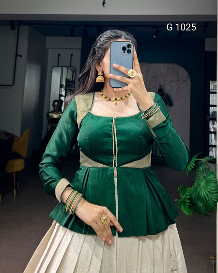 A woman takes a mirror selfie wearing a dark green traditional-style top with beige panels and a matching pleated light beige skirt. She is wearing gold jewelry and holding a blue smartphone to take the photo.