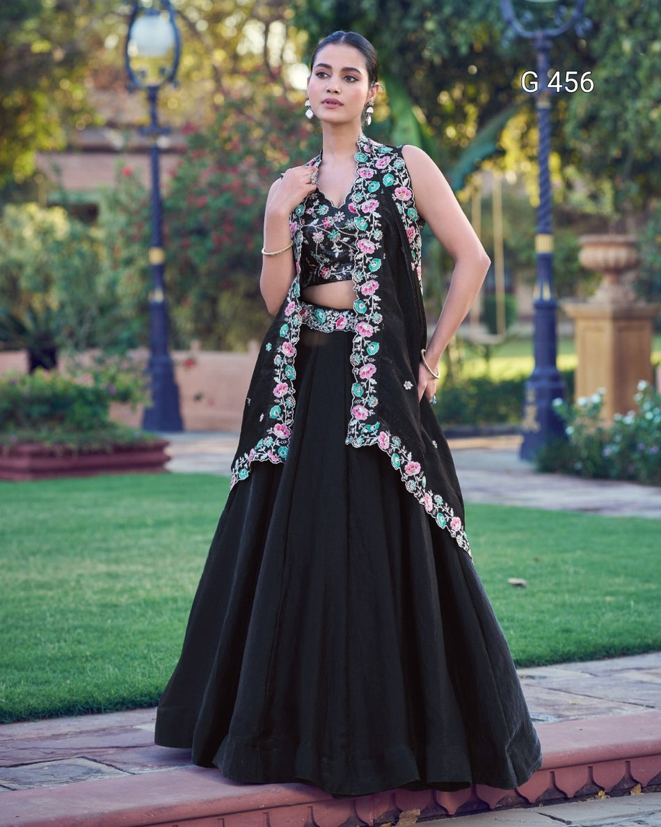 A woman stands outdoors in a garden wearing a black traditional outfit with colorful floral embroidery. The outfit includes a sleeveless draped top, a matching skirt, and a cropped waist detail.