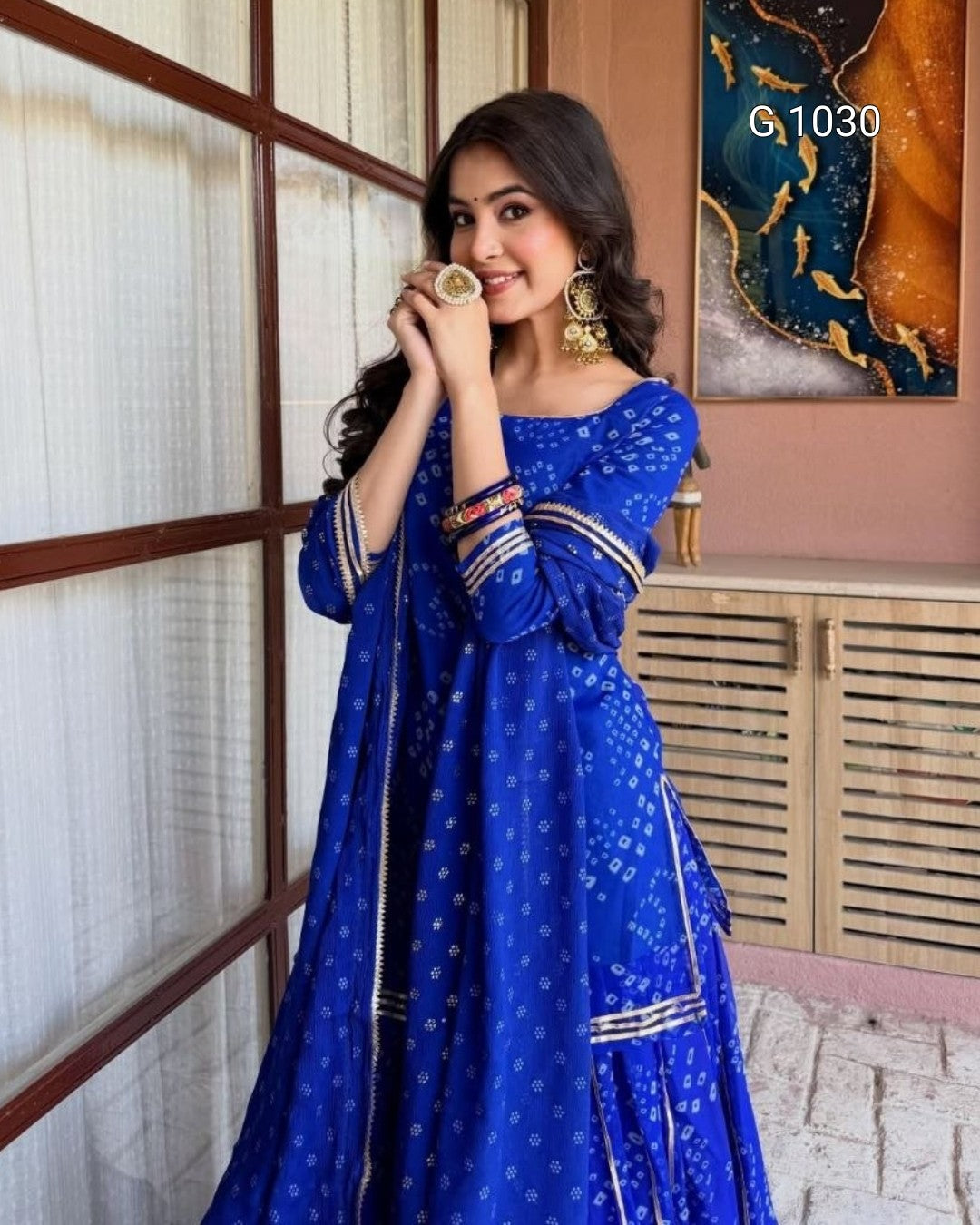 A woman wearing a royal blue Anarkali gown with white prints and gold zari embroidery, holding a small cup in front of her face. She is standing in a room with a window, wooden shutters, and a painting on the wall.