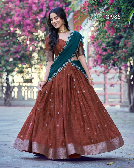 A woman wearing a rust brown embroidered lehenga choli with a dark green dupatta, standing outdoors in front of pink flowering trees. The text 'G-985' is visible in the top right corner.