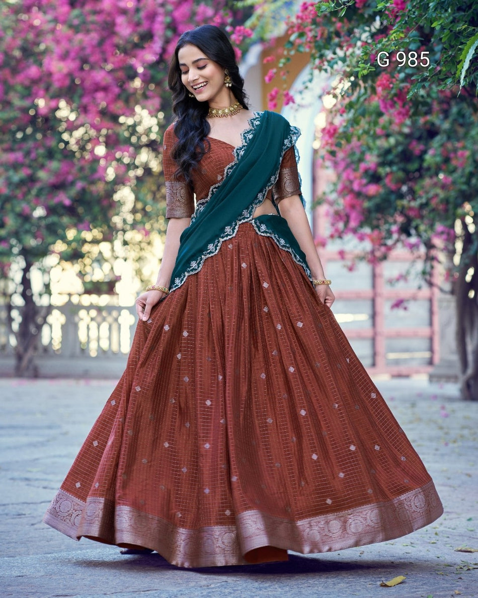 A woman wearing a rust brown embroidered lehenga choli with a dark green dupatta, standing outdoors in front of pink flowering trees. The text 'G-985' is visible in the top right corner.