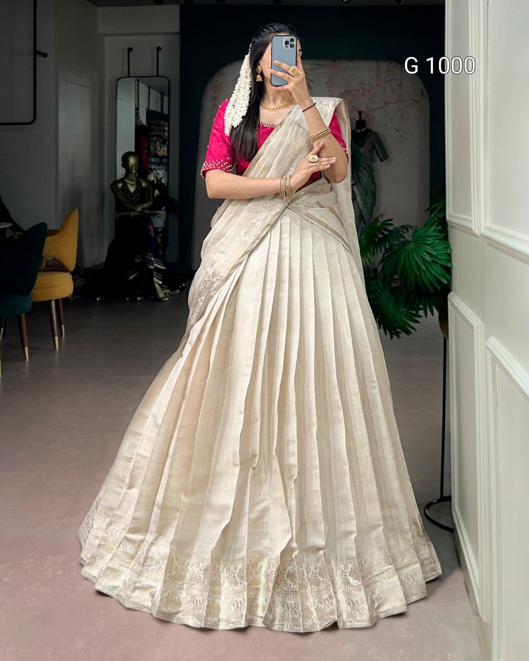 A woman takes a mirror selfie wearing a voluminous beige pleated sari with a pink blouse. The sari features subtle vertical pleats and a heavily embroidered border.