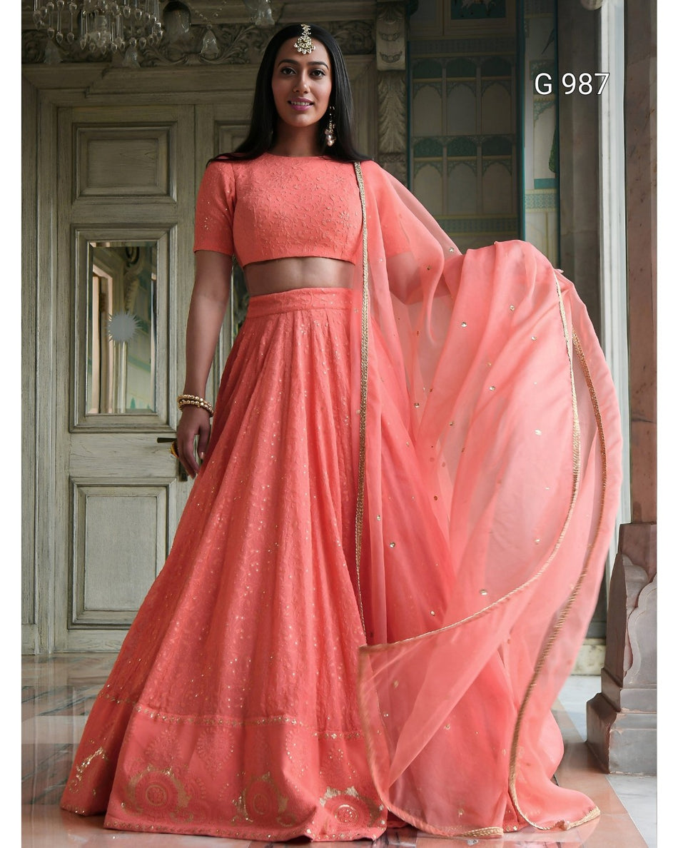 A woman models a peach-pink traditional Indian lehenga choli set with a matching dupatta in an ornate indoor setting. The set features a short-sleeved crop top, a flared skirt with subtle gold embroidery, and a sheer dupatta with a gold border.