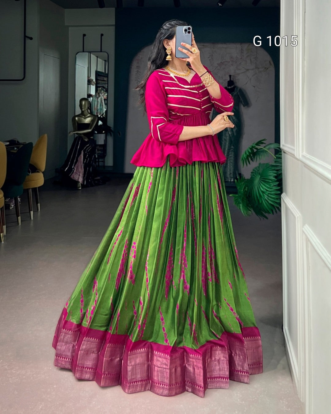 A woman takes a mirror selfie wearing a traditional Indian outfit consisting of a bright pink peplum top with white stripes and a matching green flared skirt with pink borders and vertical pink patterns.