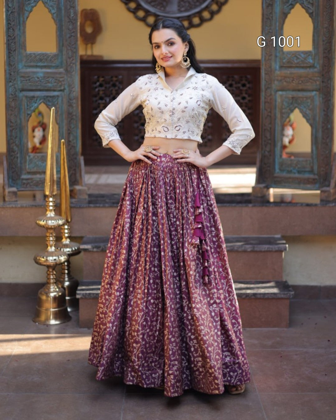 A woman models a traditional Indian ethnic wear set consisting of a white embroidered blouse and a voluminous purple skirt with gold patterns, set against an ornate courtyard background.