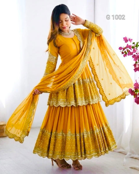 A woman modeling a traditional Indian outfit consisting of a mustard yellow anarkali-style dress with gold embroidery, a matching blouse, and a sheer dupatta, set against a bright white indoor background with pink flowers on the right.