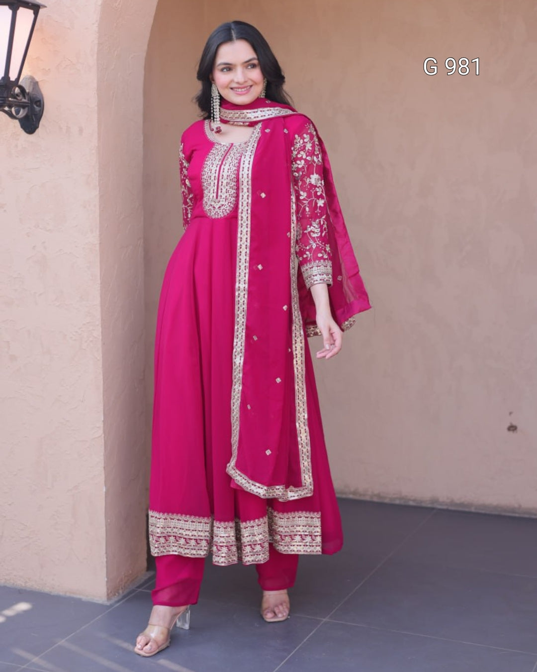 A woman models a bright magenta pink traditional Indian outfit with gold embroidery, featuring a kurta, pants, and a matching dupatta, set against a beige stucco wall background.