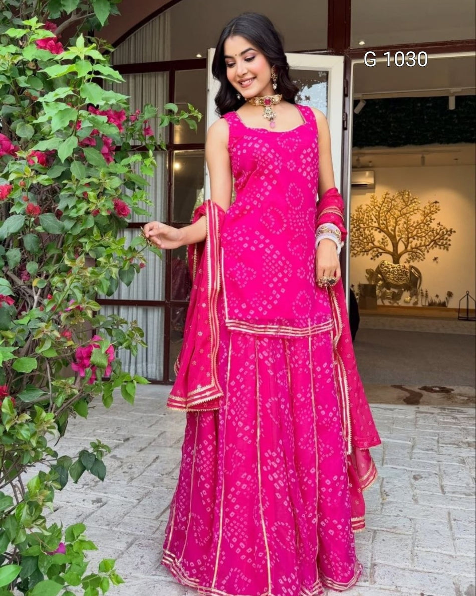 A woman modeling a bright pink Indian ethnic three-piece outfit with white floral prints and gold borders, standing outdoors next to a leafy plant. The outfit includes a sleeveless kurta, matching flared pants, and a dupatta.