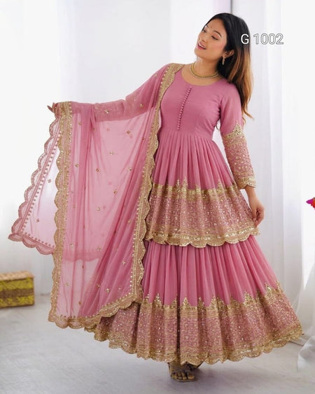 A woman modeling a pink traditional Anarkali gown with gold embroidery and a matching sheer dupatta, shown in a bright indoor setting. The text 'G1002' appears in the top right corner.