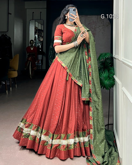 A woman takes a mirror selfie wearing a traditional Indian lehenga choli outfit. The outfit features a rust-red flared skirt (lehenga) with a decorative border, a matching rust-red blouse, and a sage green dupatta (sash) with gold motifs and a red border.