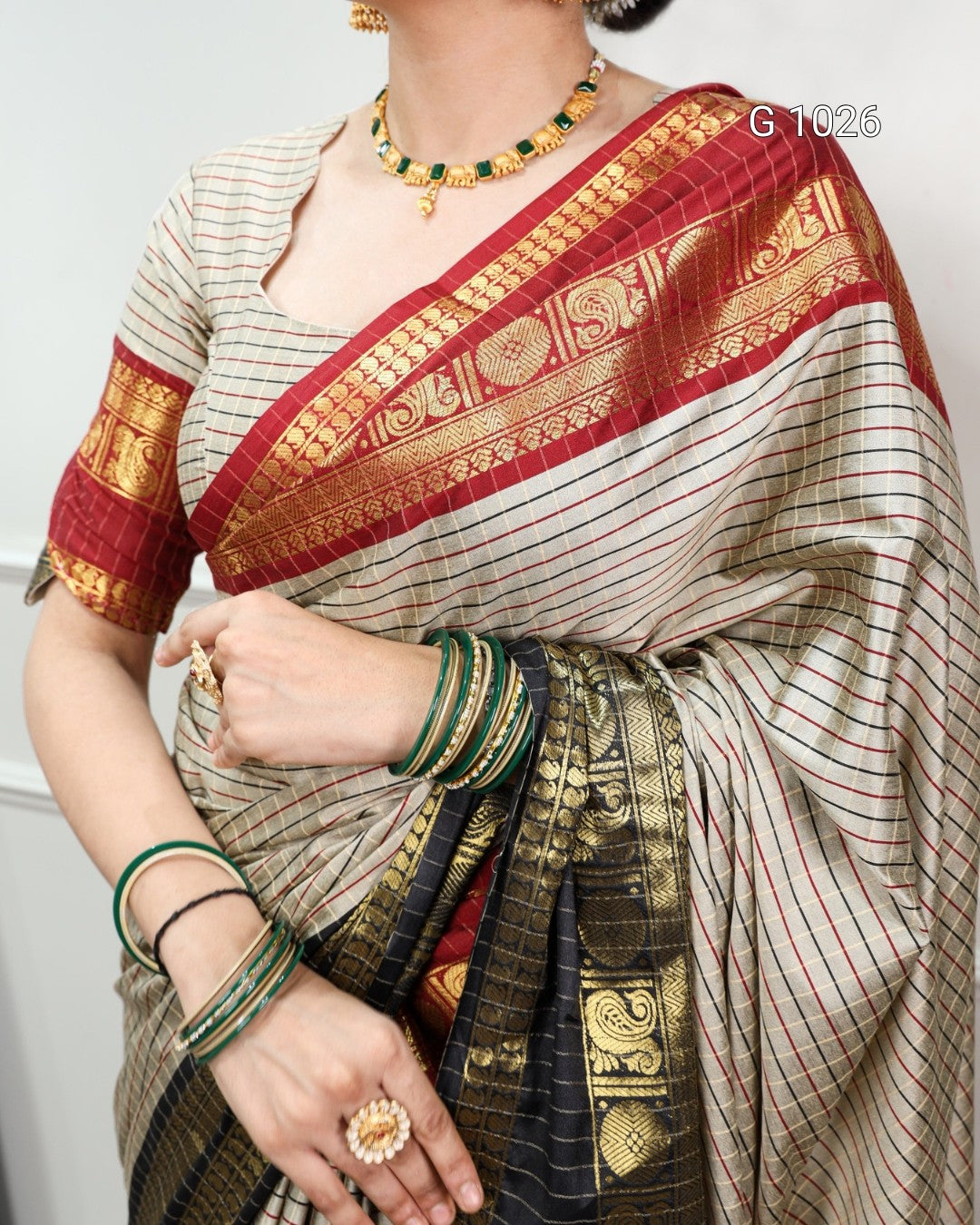 A close-up of a woman wearing a traditional Indian saree. The saree has a beige base with thin red and green stripes, a wide red border with gold zari embroidery, and a black and gold pallu.