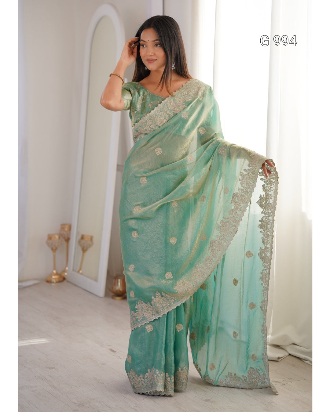 A woman models a mint green saree with gold floral motifs and a silver embroidered border, standing in a bright room with a mirror and white curtains.