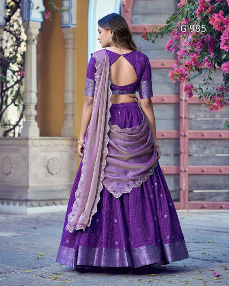 A woman wearing a purple traditional Indian outfit with a matching dupatta, shown from the back in an outdoor courtyard setting. The outfit includes a purple embroidered blouse, a purple skirt, and a light lavender dupatta draped over the shoulder.