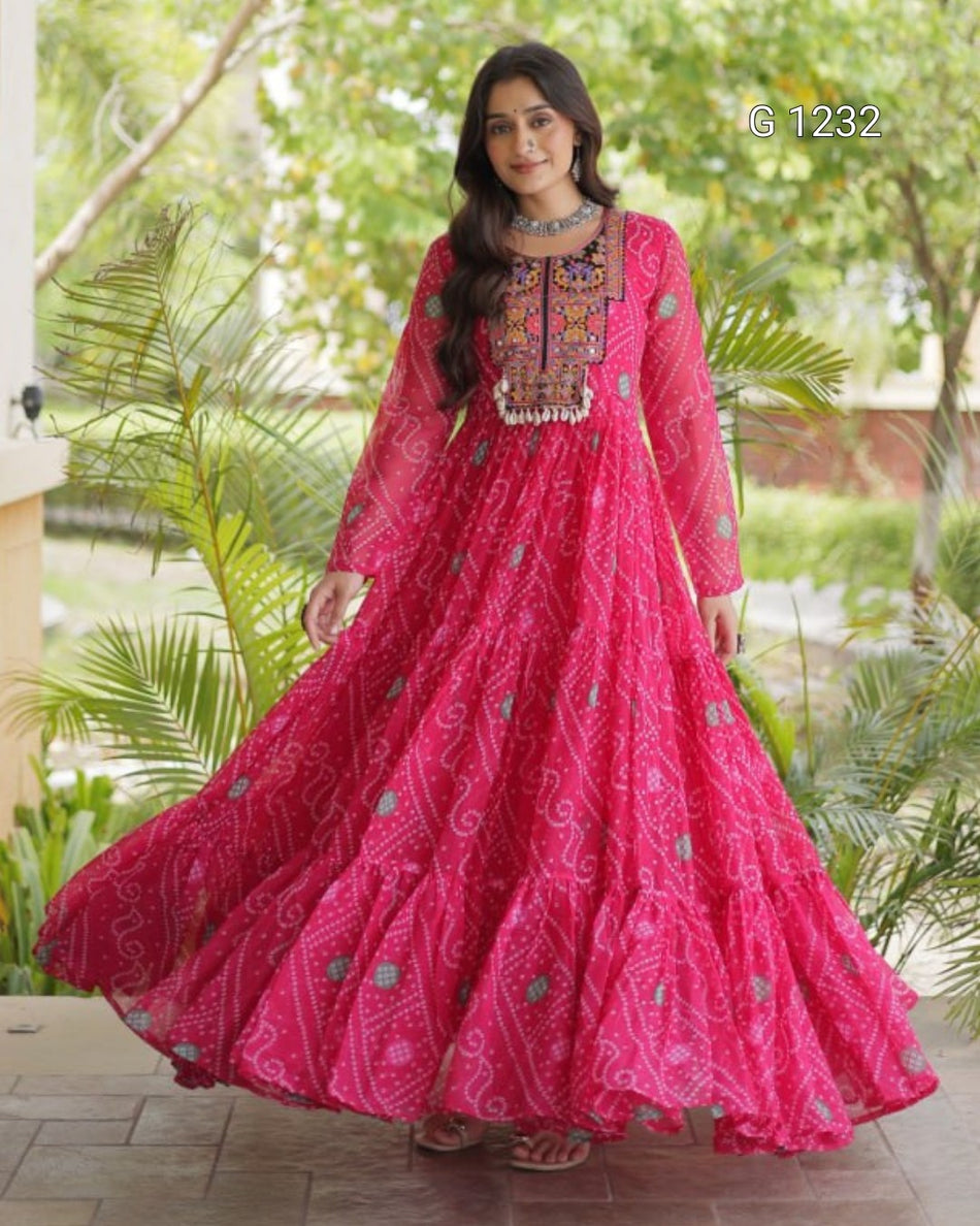 A woman wearing a bright pink traditional long dress with intricate embroidery and a flared skirt, standing outdoors in front of green foliage.