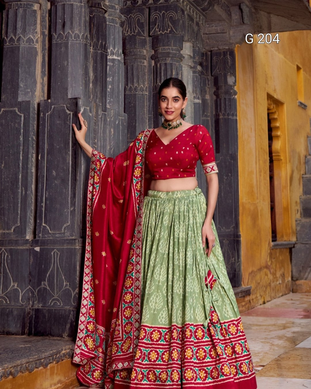 Woman in a red and green traditional outfit with a colorful dupatta against an architectural background.