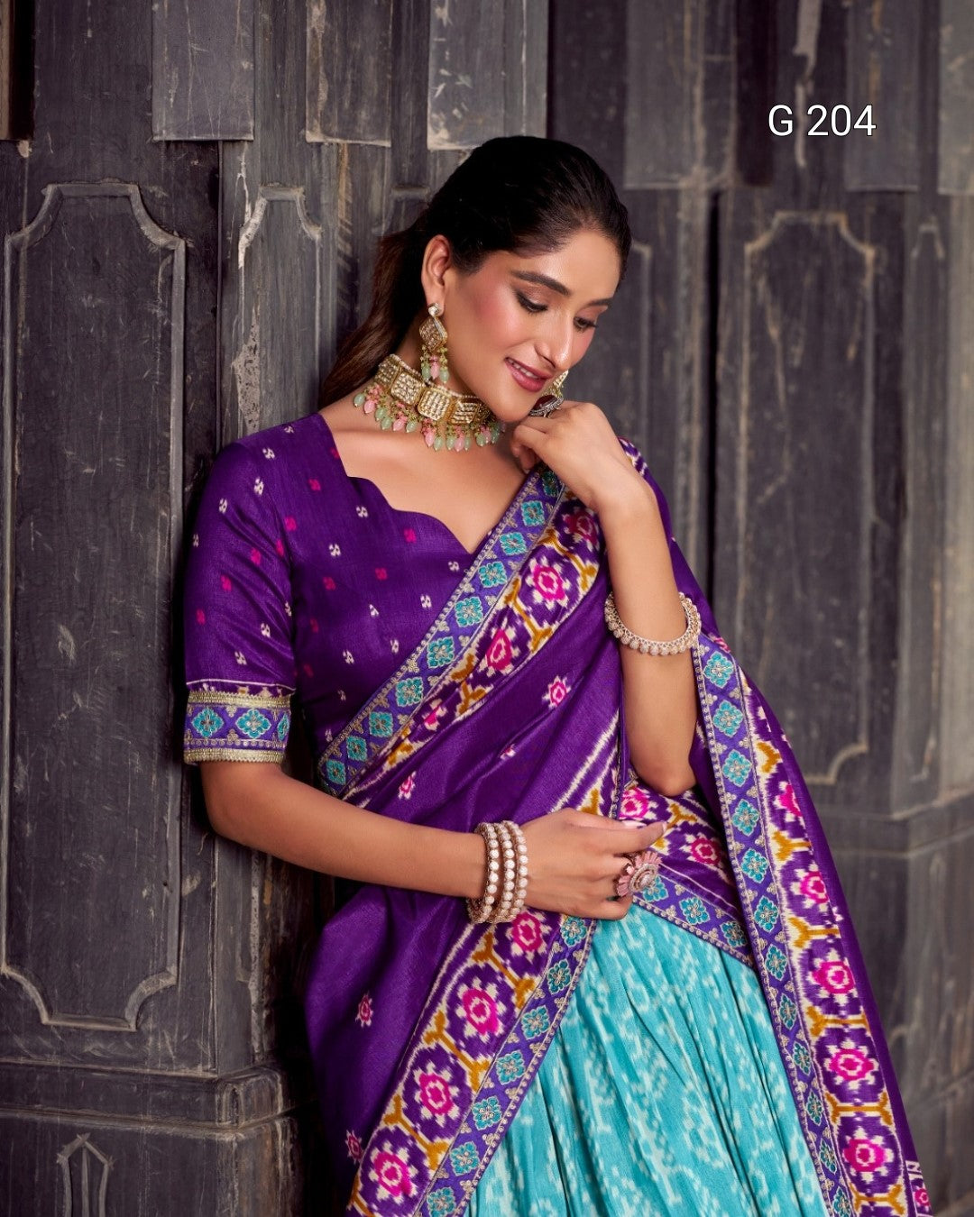 Woman wearing a purple and green saree with floral patterns against a dark stone wall.