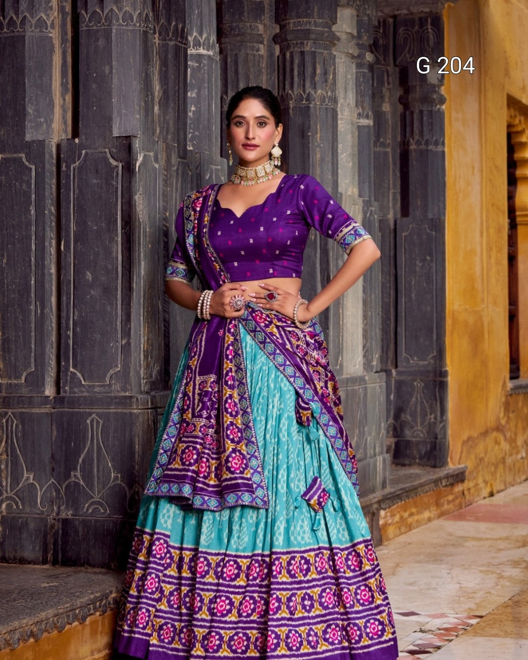 Woman in a traditional outfit with purple and teal colors standing against an architectural background.