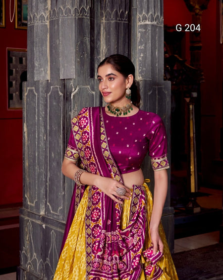 Woman in a traditional purple and yellow saree standing against a decorative wall.