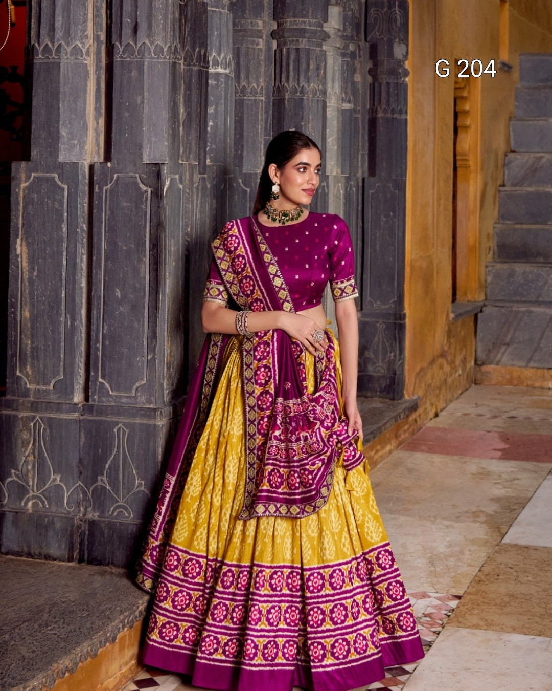Woman in a traditional yellow and purple outfit standing against a stone wall.