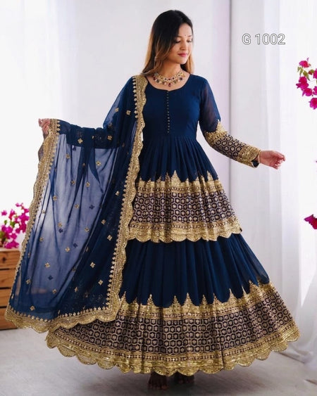 A woman modeling a navy blue traditional Anarkali gown with extensive gold embroidery and a matching sheer dupatta, set against a bright indoor background.