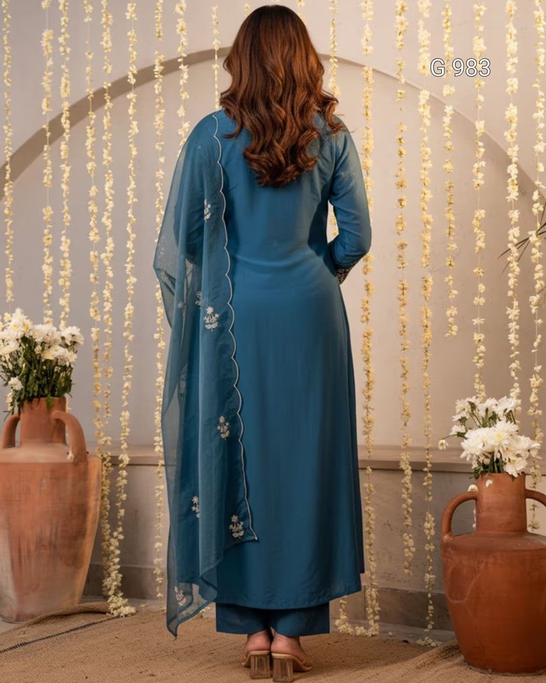 A woman models a teal blue long kurta with intricate silver embroidery on the chest, paired with matching blue trousers and a sheer blue dupatta, set against an arched backdrop decorated with hanging white flowers.