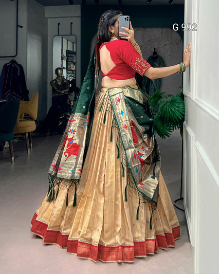 A woman wearing a traditional Indian lehenga choli outfit, taking a mirror selfie in a boutique. The outfit features a beige flared skirt (lehenga) with a red border, a red sleeveless blouse with gold embroidery, and a dark green dupatta with intricate red and gold zari embroidery.