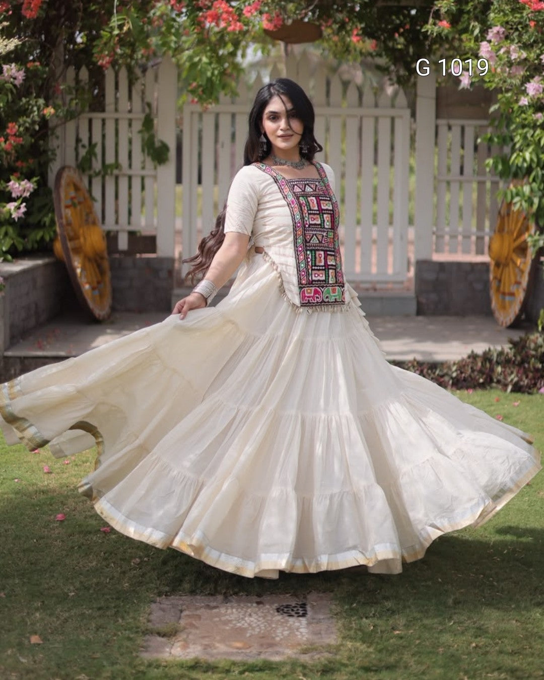 A woman outdoors wearing a cream-colored traditional Anarkali gown with a heavily embroidered multicolor chest panel. She is captured mid-twirl, causing her full skirt to flare out.