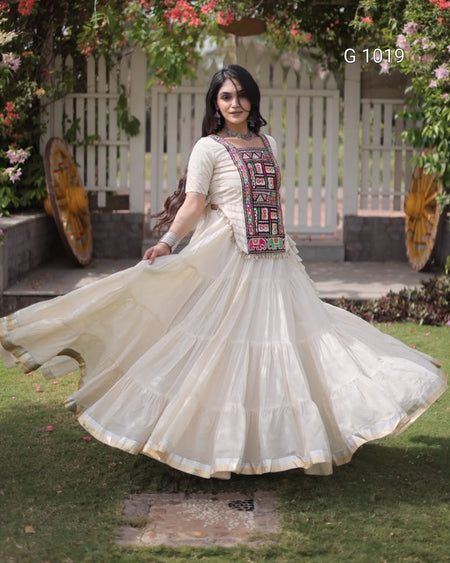 A woman outdoors wearing a cream-colored traditional Anarkali gown with a heavily embroidered multicolor chest panel. She is captured mid-twirl, causing her full skirt to flare out.